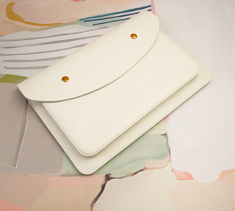 May include: A white leather envelope clutch with a flap closure and gold accents. The clutch is lying on a textured surface with abstract patterns in shades of pink, yellow, and blue.