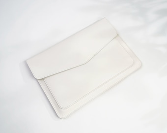 Può includere: Una custodia per laptop in pelle bianca con chiusura a lembo. La custodia è realizzata in pelle morbida e liscia e ha una lucentezza discreta.
