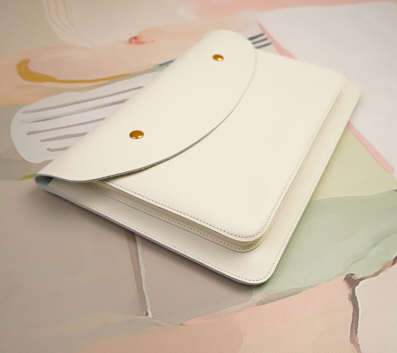 May include: A white leather notebook with a flap closure and gold accents. The notebook is lying on a light pink and green background.