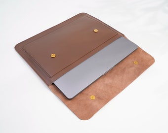 Può includere: Una custodia per laptop in pelle marrone con chiusura a lembo e bottoni a pressione dorati. La custodia è aperta e all'interno c'è un laptop.