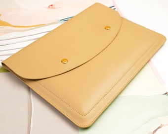 Può includere: Una custodia rettangolare per documenti o portatile di colore beige, dotata di una chiusura a patta curva fissata da due bottoni a pressione in metallo dorato. La custodia presenta cuciture tono su tono visibili lungo i bordi ben definiti.