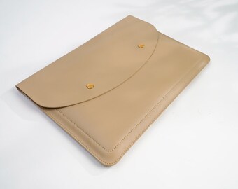 Può includere: Una custodia per laptop in pelle beige con chiusura a lembo e due bottoni a pressione in metallo dorato.