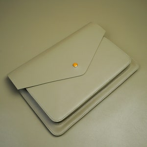 Puede incluir: Una funda para portátil de cuero gris claro de estilo sobre con cierre de botón dorado.