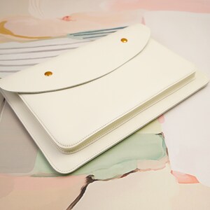 May include: A white leather envelope style clutch with a flap closure and gold metal accents. The clutch is on a light pink and green background.