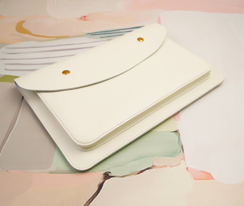 May include: A white leather envelope style clutch with a flap closure and gold metal accents. The clutch is on a light pink and green background.