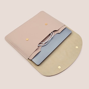 Puede incluir: Una funda para portátil de cuero color rosa pálido con solapa curva y cierres de botón a presión dorados. La funda está abierta, revelando un portátil plateado y cables de carga blancos. El interior es de color beige claro.