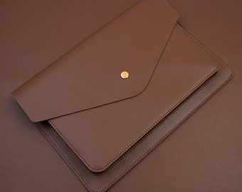 Può includere: Una custodia per laptop in pelle marrone a forma di busta con chiusura a bottone dorata.