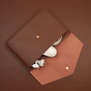 May include: A brown leather envelope-style case with a flap that opens to reveal a white cord and a white plug. The case has a small gold button on the front.