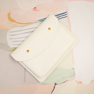 May include: A white leather envelope clutch with a flap closure and gold accents. The clutch is on a light pink and white background.