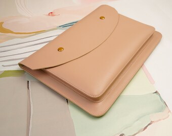 Può includere: Una custodia per laptop in pelle rosa chiaro con chiusura a lembo e dettagli in metallo dorato. La custodia ha una finitura testurizzata sottile.