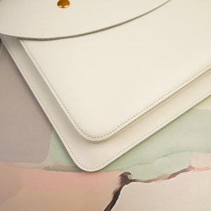 May include: A white leather laptop sleeve with a flap closure and a gold button. The sleeve is on a colorful background.