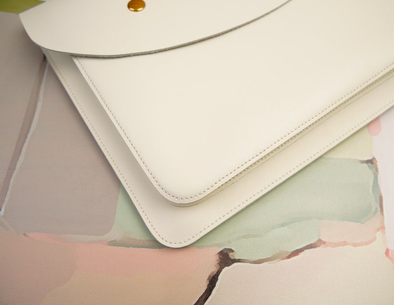 May include: A white leather laptop sleeve with a flap closure and a gold button. The sleeve is on a colorful background.