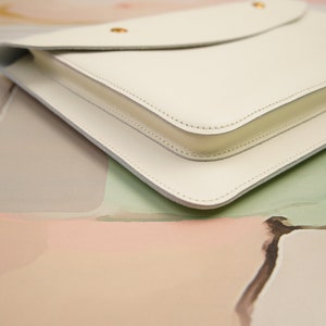 May include: A white leather clutch with a flap closure and gold hardware. The clutch is on a light pink and green background.