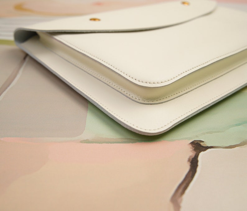 May include: A white leather clutch with a flap closure and gold hardware. The clutch is on a light pink and green background.