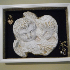 May include: White plaster cherub sculpture with gold accents, framed in a white frame with a black mat. The cherubs are embracing and have a distressed look. Two gold heart-shaped charms hang on either side of the sculpture.