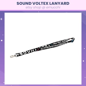 May include: Black lanyard with the text "SOUND VOLTEX" and other text in white. The lanyard has a silver-colored metal clasp. The lanyard is black with white, pink, and blue accents.