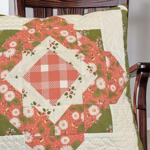 May include: A square throw pillow featuring a patchwork design with a central coral and white gingham square surrounded by floral patterns of white daisies and green leaves on coral and olive green backgrounds, finished with a white honeycomb border.
