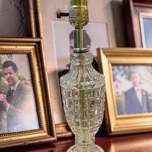 May include: A clear glass table lamp with a brass base and top. The lamp has a decorative, textured body and is set against a backdrop of framed photographs. The lamp is a vintage style.