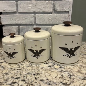 Pode incluir: Um conjunto de três potes cilíndricos em tom esbranquiçado com tampas castanho-escuras. Cada pote apresenta um gráfico castanho-escuro de uma águia com asas abertas, três pequenas estrelas acima e uma linha horizontal fina.
