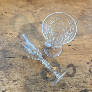 May include: Two clear, etched glass goblets on a wooden surface. One goblet is upright, showcasing the detailed floral and swirl design within the bowl. The other goblet is tilted, revealing the intricate stem design and the delicate etching.