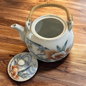 May include: White ceramic teapot with a floral design in orange, blue, and green. The teapot has a woven handle and a matching lid. The lid is off, revealing the inside of the teapot. The teapot is on a wooden surface.