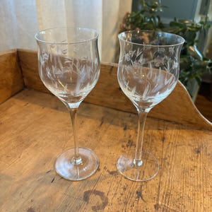 May include: Two clear wine glasses with etched floral designs. The glasses have a slender stem and a round base. The etched design features delicate flowers and leaves. The glasses are displayed on a wooden surface.