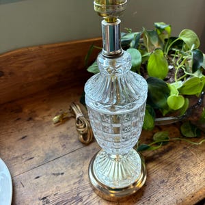 May include: A vintage table lamp with a clear, textured glass base and a gold-toned metal base and accents. The lamp has a brass-coloured socket and a brown electrical cord. The lamp is on a wooden surface.