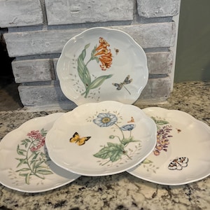 May include: A set of four white ceramic plates with scalloped edges, each featuring distinct botanical illustrations including orange flowers, blue blossoms, pink flowers, and purple clusters, accented with butterflies, bees, and dragonflies.