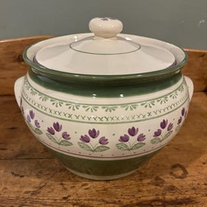 May include: A ceramic lidded serving bowl with a floral design. The bowl is white with green and purple accents. The lid has a small knob. The bowl has two handles and a decorative band of purple flowers.