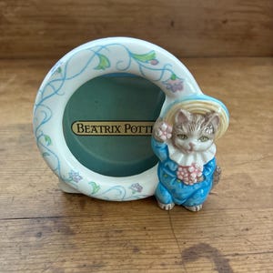 May include: A ceramic picture frame with a light blue and white floral design. The frame has a round opening with the text "BEATRIX POTTER" and features a figurine of a cat wearing a blue outfit and hat.