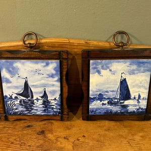 May include: Two framed ceramic tiles with a nautical theme. Each tile features a blue and white sailboat scene with a dark wood frame and a brass ring for hanging. The artwork depicts sailboats on a body of water.
