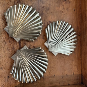 Pode incluir: Três objetos decorativos prateados em forma de concha. Cada concha tem um design em forma de leque com nervuras verticais e uma base retangular. Os objetos estão dispostos sobre uma superfície de madeira, criando um tema costeiro ou náutico.