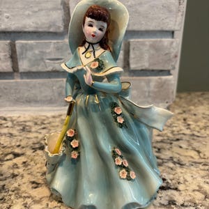 Può includere: Una statuetta in porcellana raffigurante una persona con un abito azzurro con finiture dorate e piccoli accenti floreali rosa. La figura indossa un cappello a tesa larga, un girocollo nero con ciondolo dorato e tiene un ombrellino giallo.