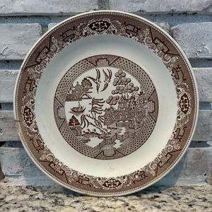 May include: A round ceramic plate featuring a brown transferware pattern on a cream background. The central design depicts a landscape with a pagoda, a bridge, a boat, and two birds. The rim is decorated with a geometric and floral border.