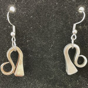 Puede incluir: Un par de pendientes de plata con forma del signo astrológico Leo. Los pendientes están hechos de metal y tienen un diseño simple y elegante.