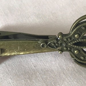 Puede incluir: Un pasador de corbata de color latón antiguo con un diseño ornamental y decorativo. El pasador presenta un patrón calado detallado con pequeños puntos en relieve y un acento floral. El pasador está diseñado para asegurar una corbata.