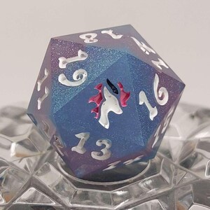 May include: A twenty-sided die featuring a gradient of blue and purple hues with a shimmering, glittery finish. The numbers are engraved in white, and a small, stylized white and red unicorn graphic is positioned near the number sixteen.