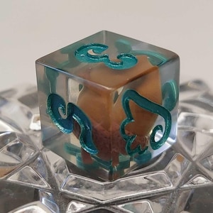 Op de afbeelding: Een doorzichtige harsdobbelsteen met turquoise wervelingen en een bruine kern. De kubusvormige dobbelsteen heeft turquoise ontwerpen en ligt op een facetgeslepen glazen oppervlak. De turquoise ontwerpen zijn aan meerdere zijden zichtbaar.