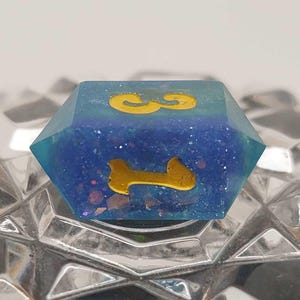 Puede incluir: Un dado hexagonal de resina azul con los números dorados 1 y 3. El dado está lleno de purpurina y pequeñas motas de colores. El dado está sobre una superficie plateada reflectante.