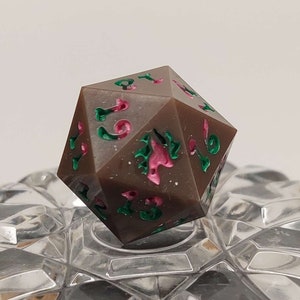 Op de afbeelding: Een bruine, veelzijdige dobbelsteen met roze en groene accenten. De dobbelsteen ligt op een helder glazen oppervlak.