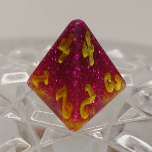 May include: A pink and yellow glittery d4 dice with gold numbers. The dice is a four-sided pyramid shape.