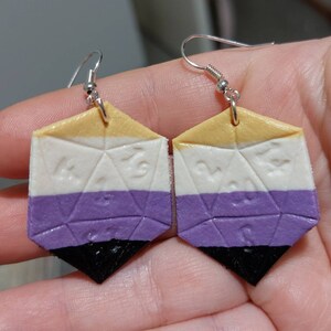 May include: A pair of dangle earrings shaped like a 20-sided die, painted in the colors of the non-binary pride flag: yellow, white, purple, and black.