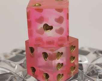 May include: Two pink dice with gold heart accents stacked on top of each other. The dice are made of a translucent material and sit on a decorative glass surface. The dice are a fun and unique item.
