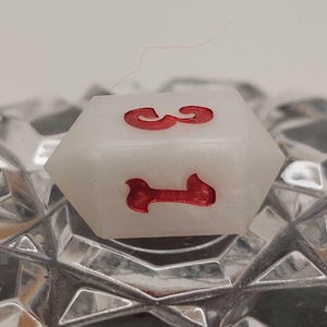 May include: A white, multi-sided die with red numbers 1 and 3. The die is resting on a textured, clear surface. The die is a game piece, likely for a tabletop game.