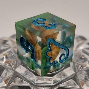 May include: A clear resin die with teal-colored numbers and gold-colored inclusions. The die is cube-shaped and sits on a textured surface. The numbers are stylized and the die has a green tint.
