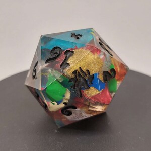 Può includere: Un dado di resina trasparente multicolore con numeri neri e un design nero. Il dado presenta una varietà di colori, tra cui rosso, blu, verde, oro e bianco.