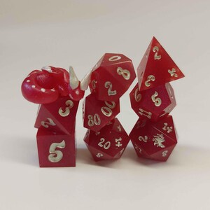 May include: A set of seven translucent red polyhedral dice stacked in three columns. The dice feature silver-colored numbers and a small, sculpted red dragon with white accents resting on top of the leftmost die. The set includes various shapes.