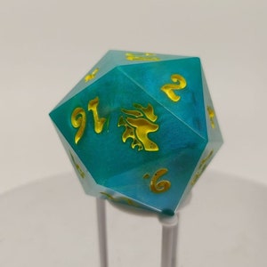 Könnte beinhalten: Ein türkisfarbener, schimmernder 20-seitiger Würfel mit goldenen Zahlen und einem Drachenmotiv. Der Würfel steht auf einem weißen Ständer.