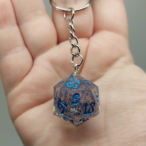 Può includere: Un portachiavi a forma di dado a venti facce con un corpo in resina traslucida riempito di inclusioni scure, glitterate e metalliche. Il dado presenta numeri dipinti in blu brillante ed è fissato a un anello in metallo argentato tramite una catenella.