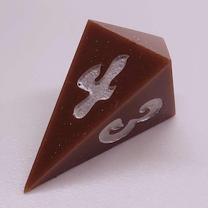 May include: A brown, triangular-shaped dice with silver-colored, stylized markings. The dice has a smooth, polished surface and is set against a plain white background. The markings appear to be decorative.